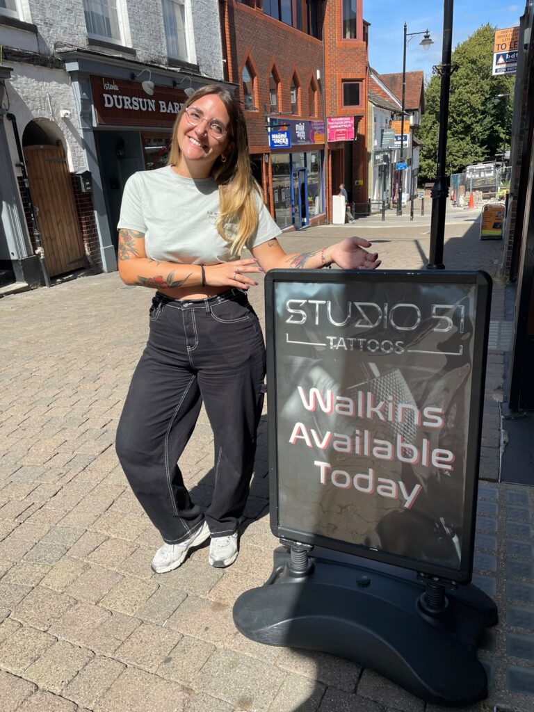 Smiling woman with tattoos standing on a sunny street beside a Studio 51 Tattoos sign offering walk-ins today.